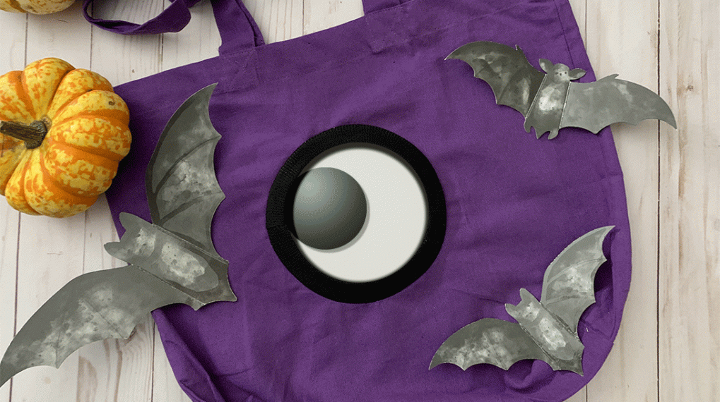 An animated flat lay scene featuring a purple tote bag with a giant googley eye that shifts position every two seconds. Surrounding the bag are paper bats and a few pumpkins.