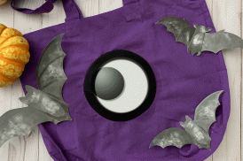 An animated flat lay scene featuring a purple tote bag with a giant googley eye that shifts position every two seconds. Surrounding the bag are paper bats and a few pumpkins.