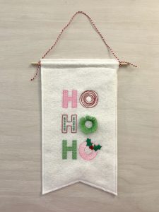 Machine embroidered wall hanging with the words, "Ho, Ho, Ho" written in stylized letters using a variety of stitches in red, pink, light green, and dark green.