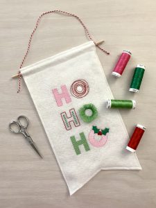 Small felt banner featuring embroidered letters spelling, "Ho! Ho! Ho!" The middle "O" is a special "fringe" stitch that adds a 3D effect that resembles a wreath when the bobbin threads are cut from the back side.