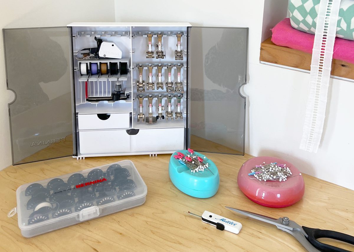 An array of sewing machine accessories, including a small box of bobbins, a case full of sewing machine feet, two pincushions, and scissors.