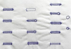 A grid of 13 buttonholes and 2 eyelets sewn with purple thread on white fabric.