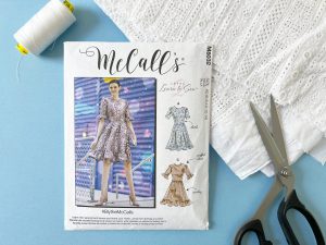 Flatly featuring a white spool of thread, McCall's sewing pattern with a woman wearing a dress on the cover, white eyelet fabric and a pair of scissors.