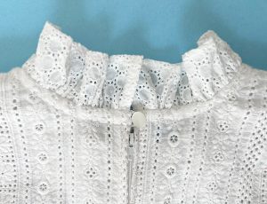 Back neckline of a white eyelet dress, featuring a ruffle, a small white button, and a centered zipper.