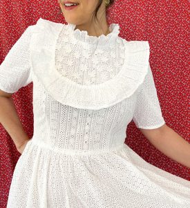 A woman's torso wearing a white eyelet dress with short sleeves, a high ruffled neckline, bodice ruffle and appliqué flowers.