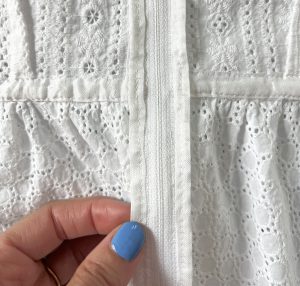 White eyelet fabric with a zipper going through the center of the image. The sides of the zipper tape have been bound is white rayon ribbon. A hand with blue nail polish is pinching the binding.
