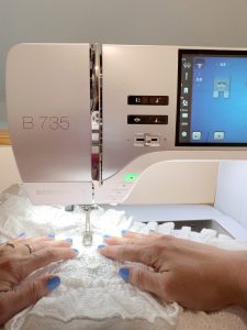 A sewing machine with white fabric under the needle. Two hands are guiding the fabric.