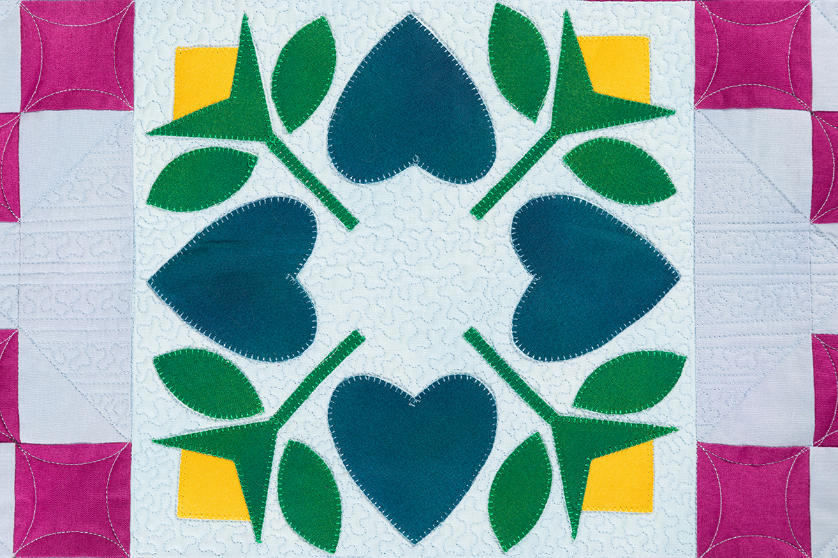 The Heart & Flower Applique Block shown in blue, green, yellow, and pink, on a white background.