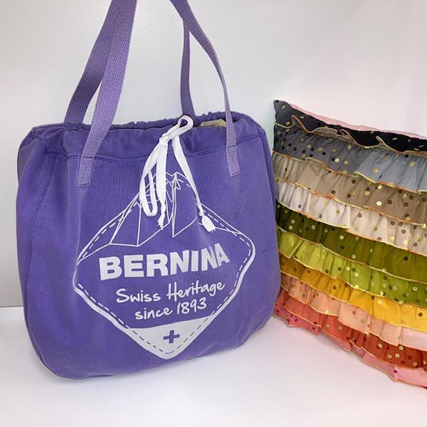 Purple BERNINA tote bag next to rainbow ruffled pillow on white background