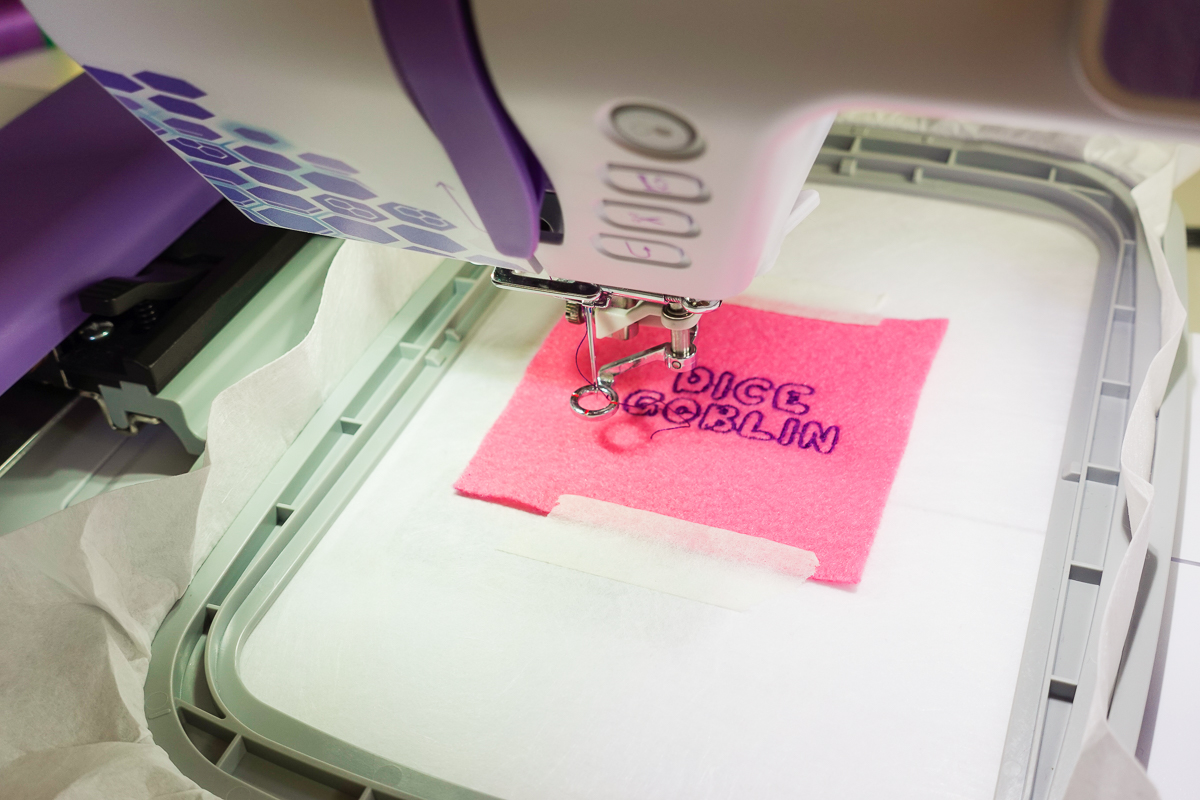 embroidery in progress of purple thread lettering dice goblin on pink felt