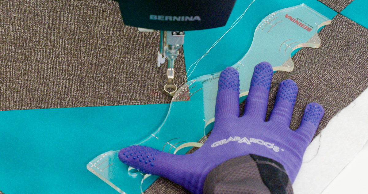 Close-up photo of purple-gloved hands quilting curvy lines on teal and gray fabric with a BERNINA Ruler.