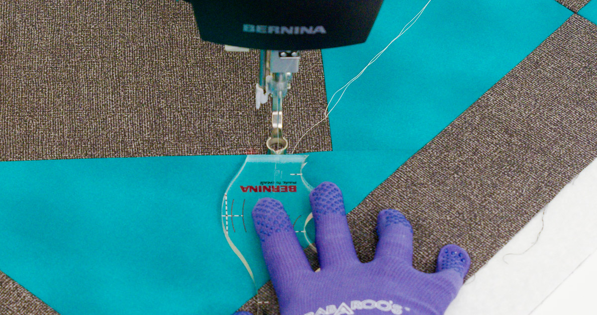 Close-up photo of purple-gloved hands quilting curvy lines on teal and gray fabric with a BERNINA Ruler.