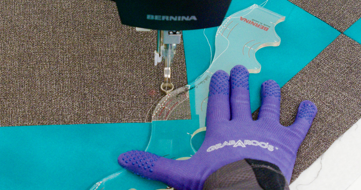 Close-up photo of purple-gloved hands quilting curvy lines on teal and gray fabric with a BERNINA Ruler.