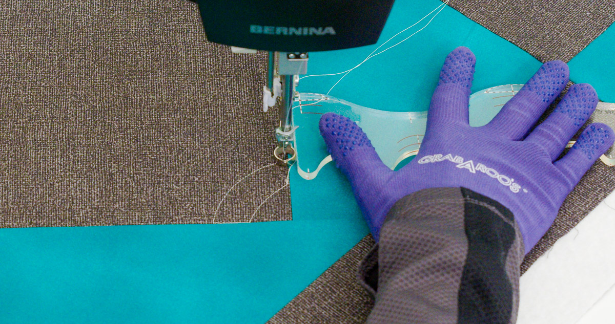 Close-up photo of purple-gloved hands quilting curvy lines on teal and gray fabric with a BERNINA Ruler.