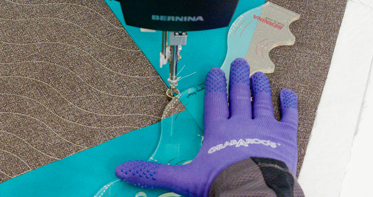 Close-up photo of purple-gloved hands quilting curvy lines on teal and gray fabric with a BERNINA Ruler.