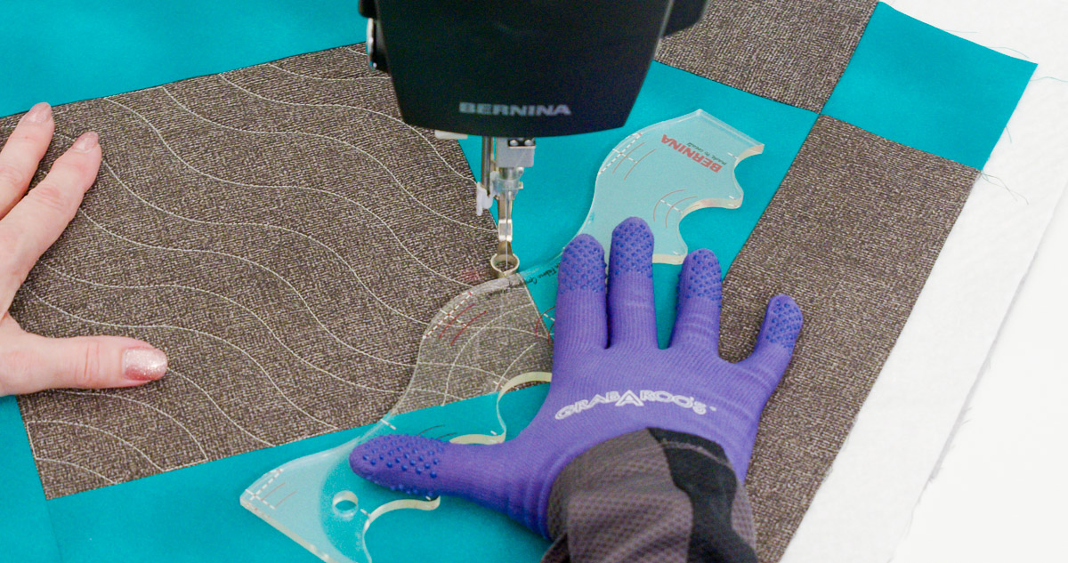 Close-up photo of purple-gloved hands quilting curvy lines on teal and gray fabric with a BERNINA Ruler.