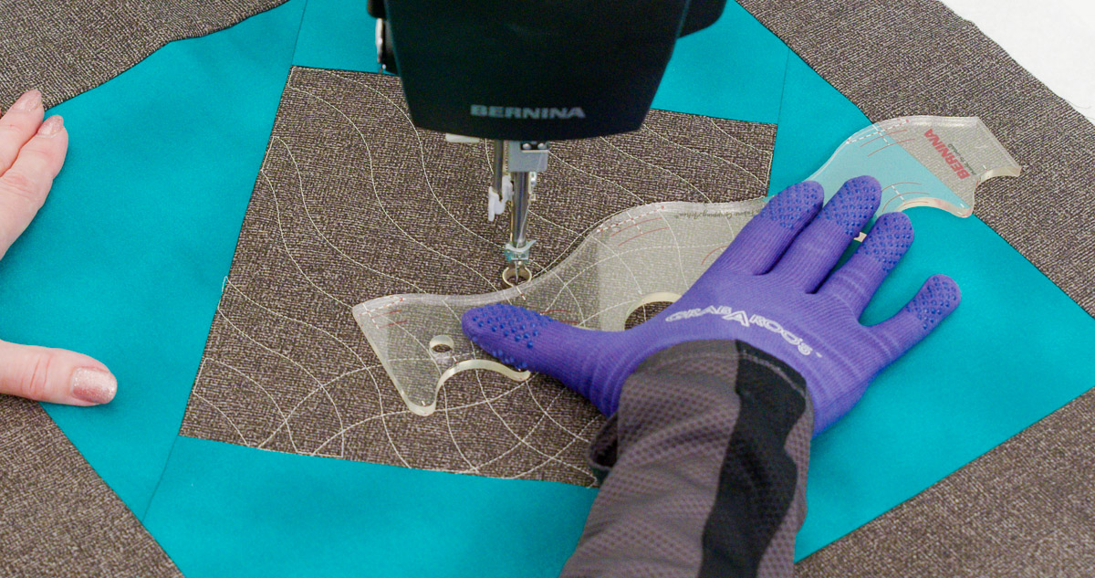 Close-up photo of purple-gloved hands quilting curvy lines on teal and gray fabric with a BERNINA Ruler.