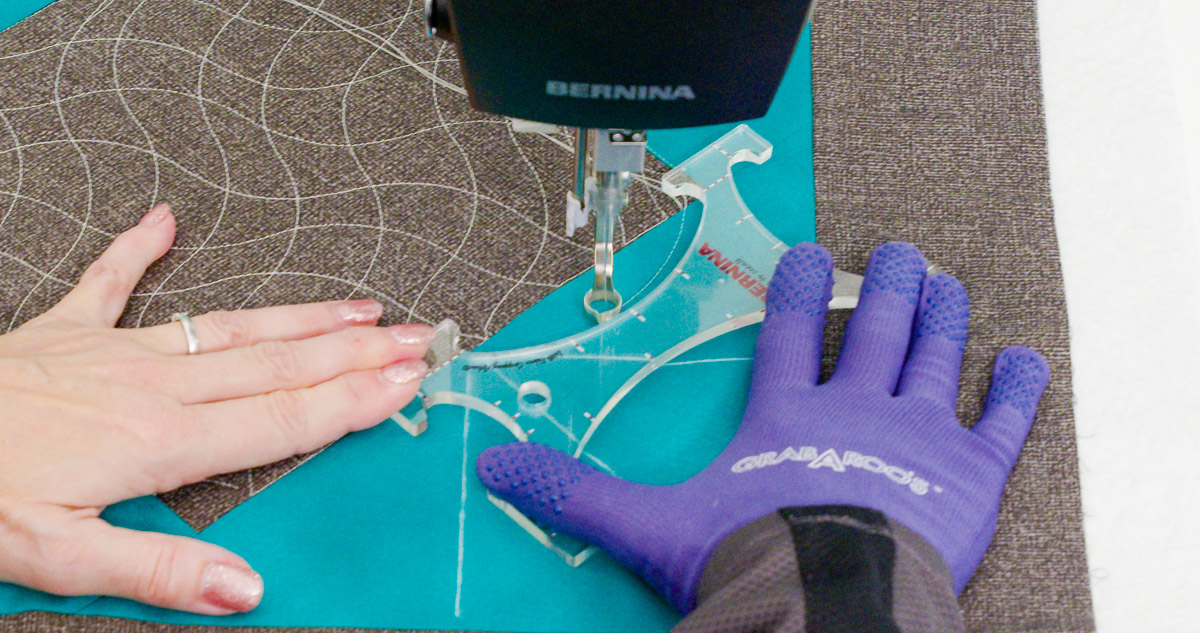 Close-up photo of purple-gloved hands quilting curvy lines on teal and gray fabric with a BERNINA Ruler.