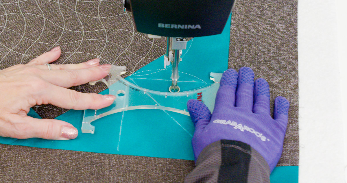 Close-up photo of purple-gloved hands quilting curvy lines on teal and gray fabric with a BERNINA Ruler.