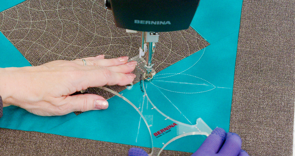 Close-up photo of purple-gloved hands quilting curvy lines on teal and gray fabric with a BERNINA Ruler.