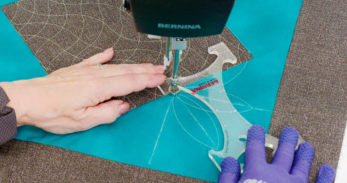 Close-up photo of purple-gloved hands quilting curvy lines on teal and gray fabric with a BERNINA Ruler.