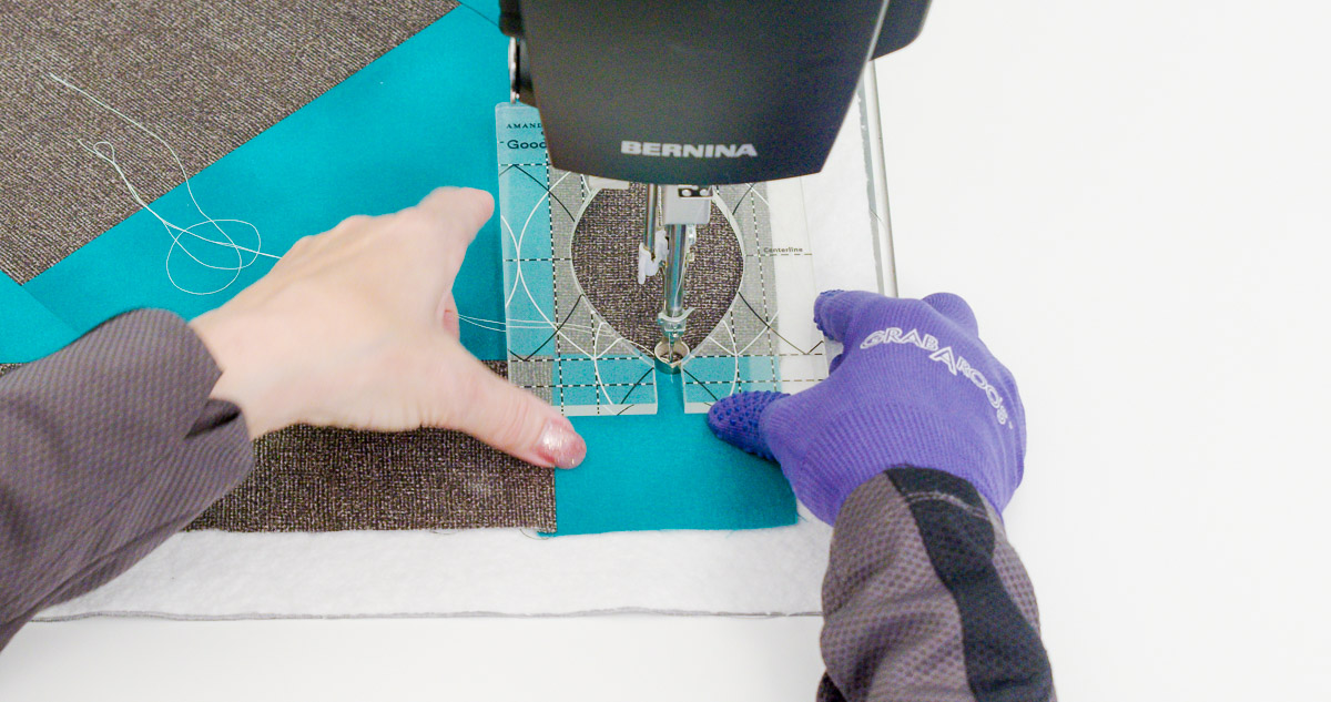 Close-up photo of purple-gloved hands quilting curvy lines on teal and gray fabric with a Good Measure Leaf Ruler from Amanda Murphy.
