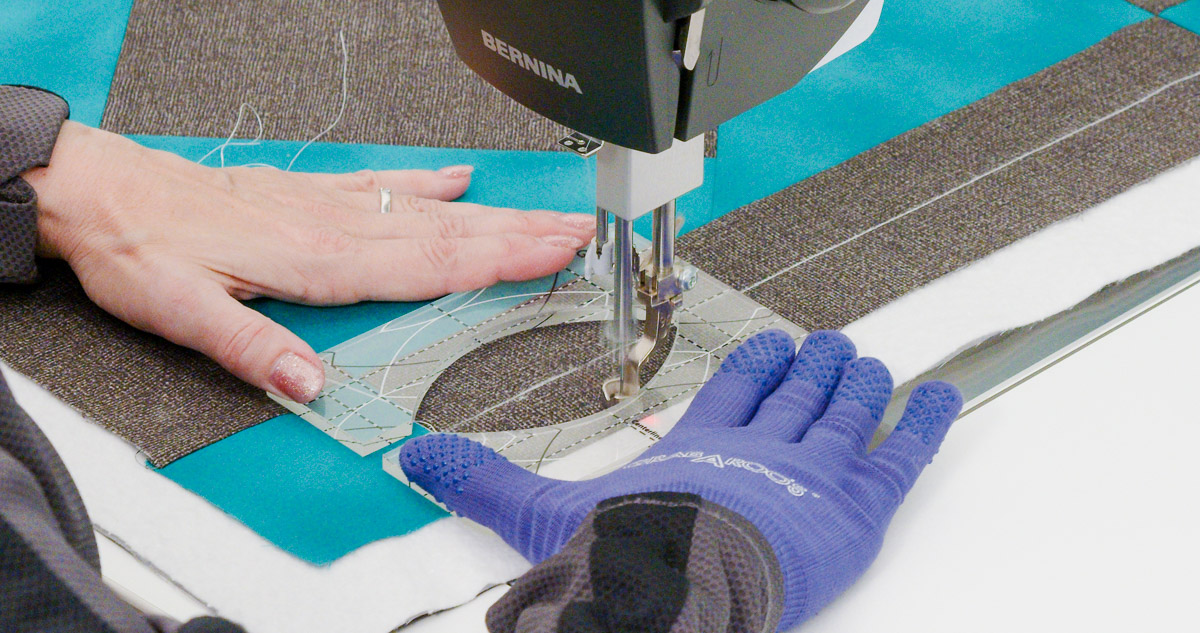 Close-up photo of purple-gloved hands quilting curvy lines on teal and gray fabric with a Good Measure Leaf Ruler from Amanda Murphy.