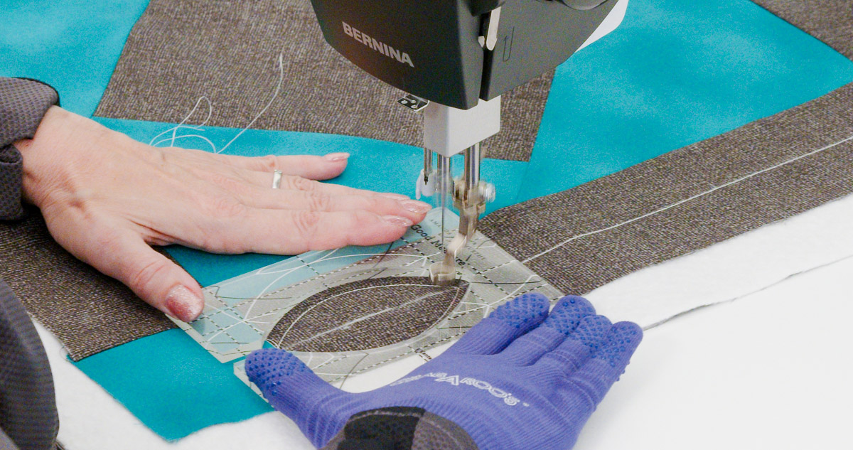 Close-up photo of purple-gloved hands quilting curvy lines on teal and gray fabric with a Good Measure Leaf Ruler from Amanda Murphy.