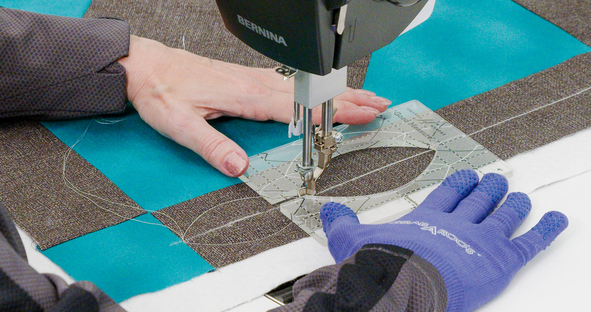 Close-up photo of purple-gloved hands quilting curvy lines on teal and gray fabric with a Good Measure Leaf Ruler from Amanda Murphy.