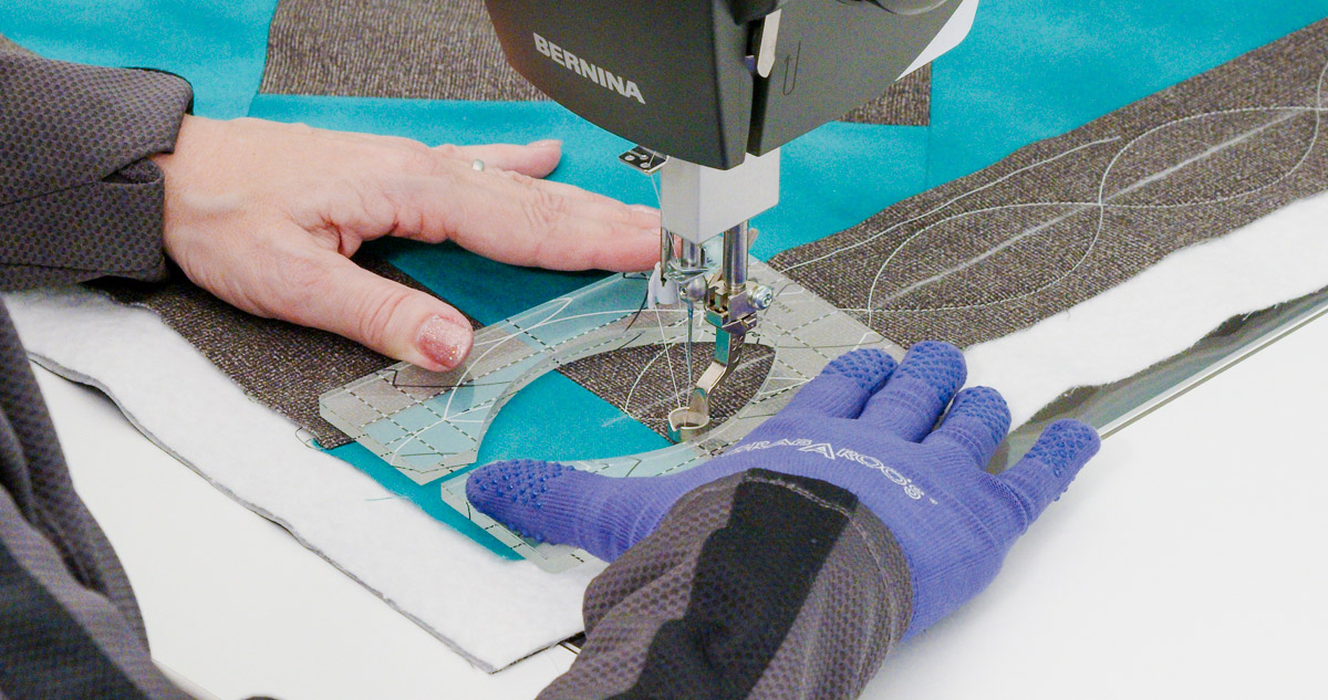 Close-up photo of purple-gloved hands quilting curvy lines on teal and gray fabric with a Good Measure Leaf Ruler from Amanda Murphy.