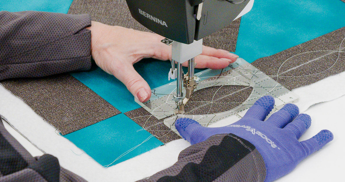 Close-up photo of purple-gloved hands quilting curvy lines on teal and gray fabric with a Good Measure Leaf Ruler from Amanda Murphy.