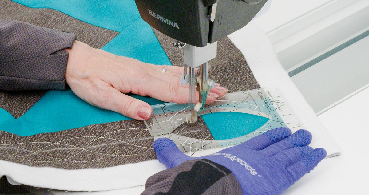 Close-up photo of purple-gloved hands quilting curvy lines on teal and gray fabric with a Good Measure Leaf Ruler from Amanda Murphy.