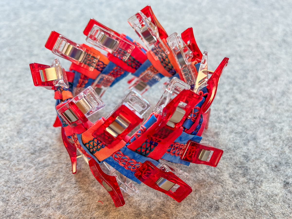 finished serger cuff bracelet in orange and blue fabric, covered in red clips, on a gray background
