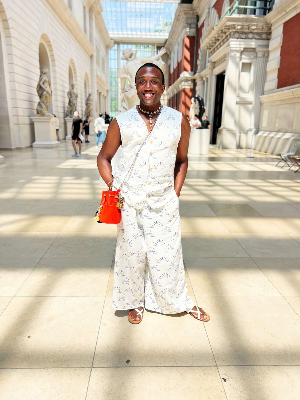 A person standing in a bright museum atrium wearing a matching sleeveless button-front top and wide-leg pants made from white fabric with a blue and yellow abstract print. They are smiling, holding a small bright orange bag, and wearing sandals. Marble statues and tall windows fill the background.