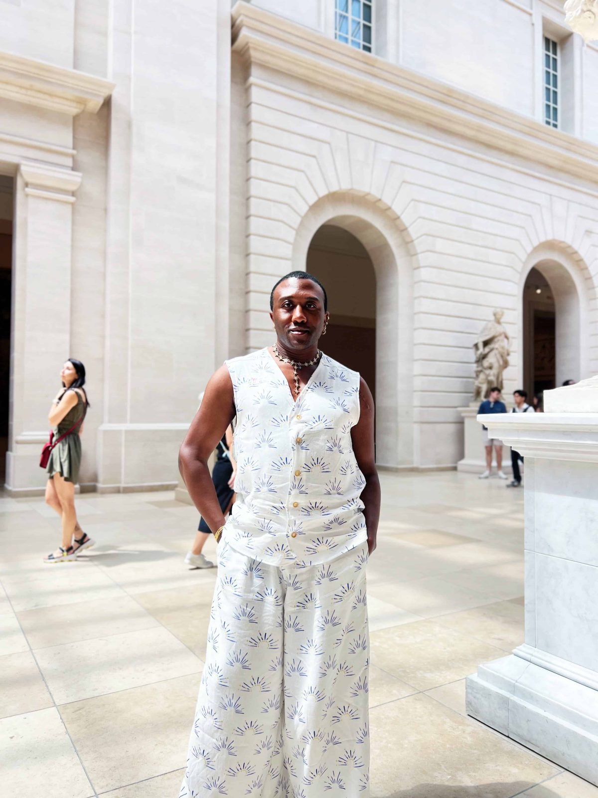 A person standing in a bright museum atrium wearing a matching sleeveless button-front top and wide-leg pants made from white fabric with a blue and yellow sun print. They are smiling, holding a small bright orange bag, and wearing sandals. Marble statues and tall windows fill the background.