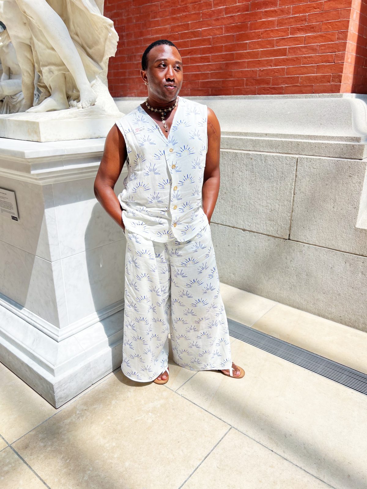 A person stands in a museum beside a marble statue, wearing a sleeveless button-front top and wide-leg pants made from white fabric with a blue and yellow sun print. They have their hands in their pockets and are looking slightly to the side, with sunlight casting soft shadows on the floor.