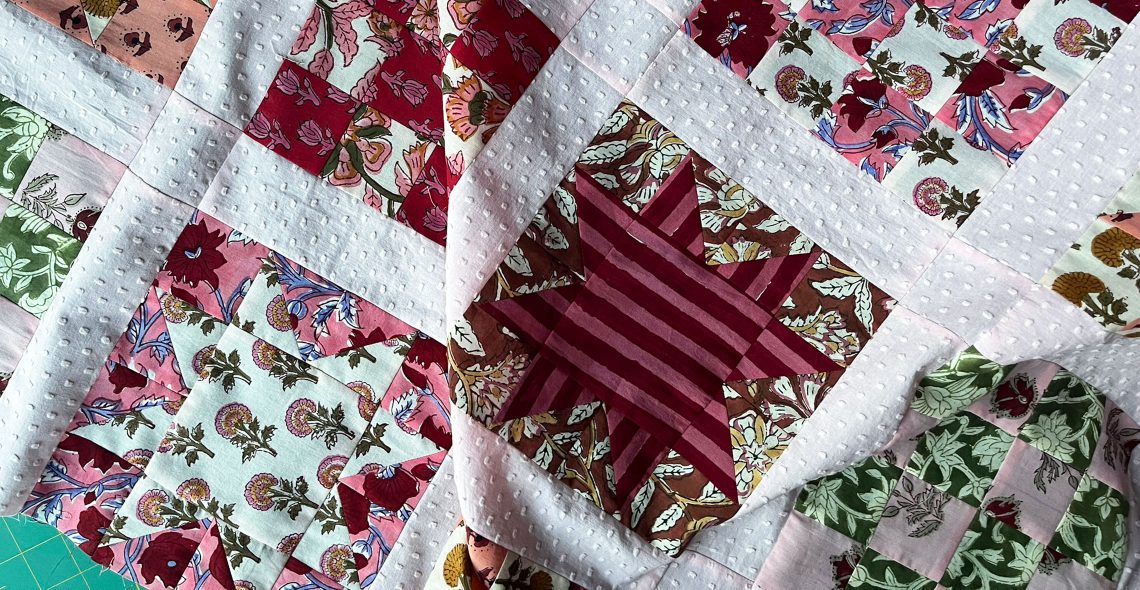 A close up of a quilt in progress. The quilt features block prints arranged in alternating quilt blocks of 16-patches and sawtooth stars. The colors are mostly in shades of pink and red.