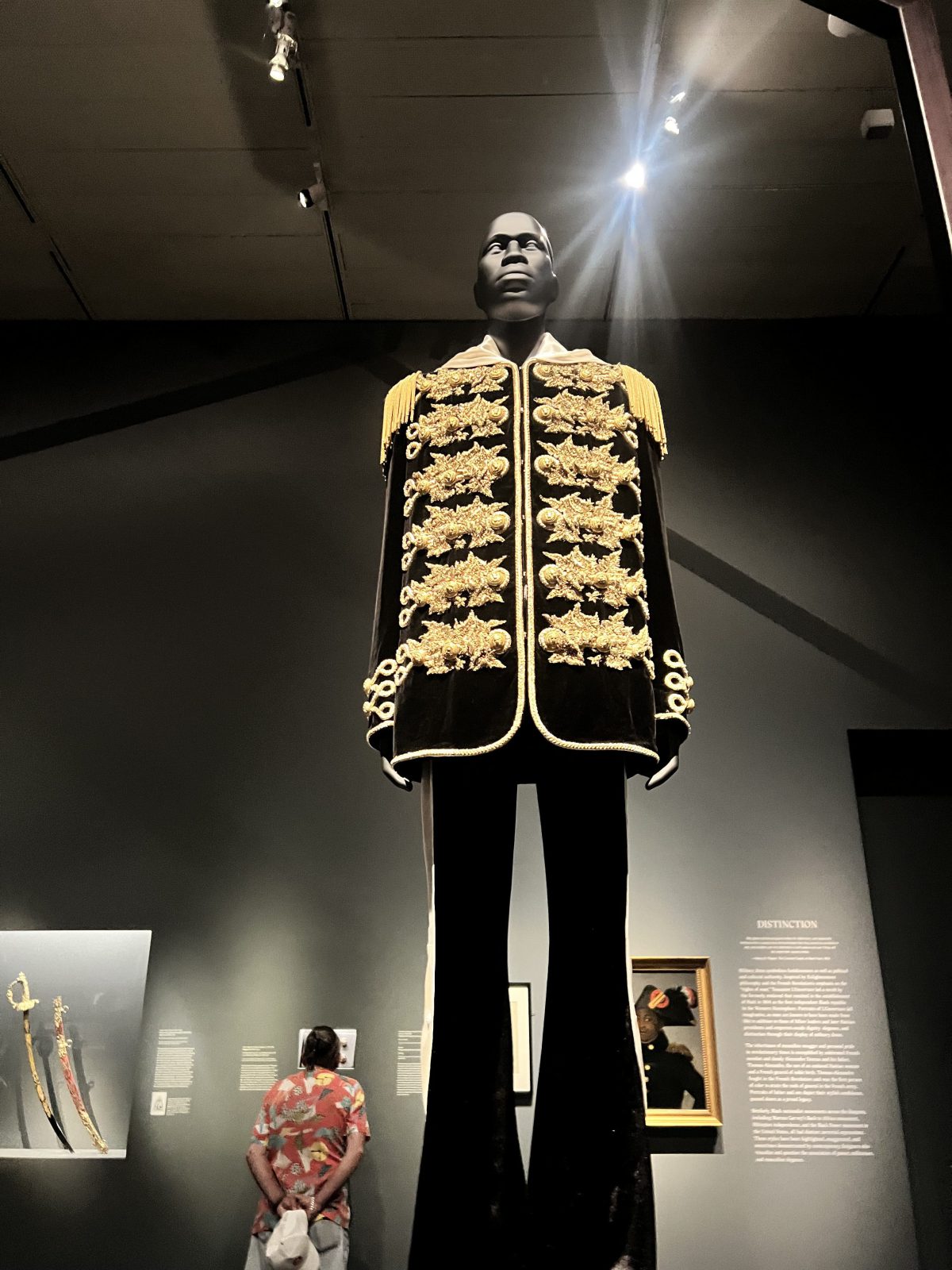 A tall mannequin wearing a black velvet military-style jacket adorned with elaborate gold embroidery and shoulder epaulettes stands in a museum gallery. A visitor in a colorful shirt stands below, looking up at the display. Dim gallery lighting highlights the garment’s detail