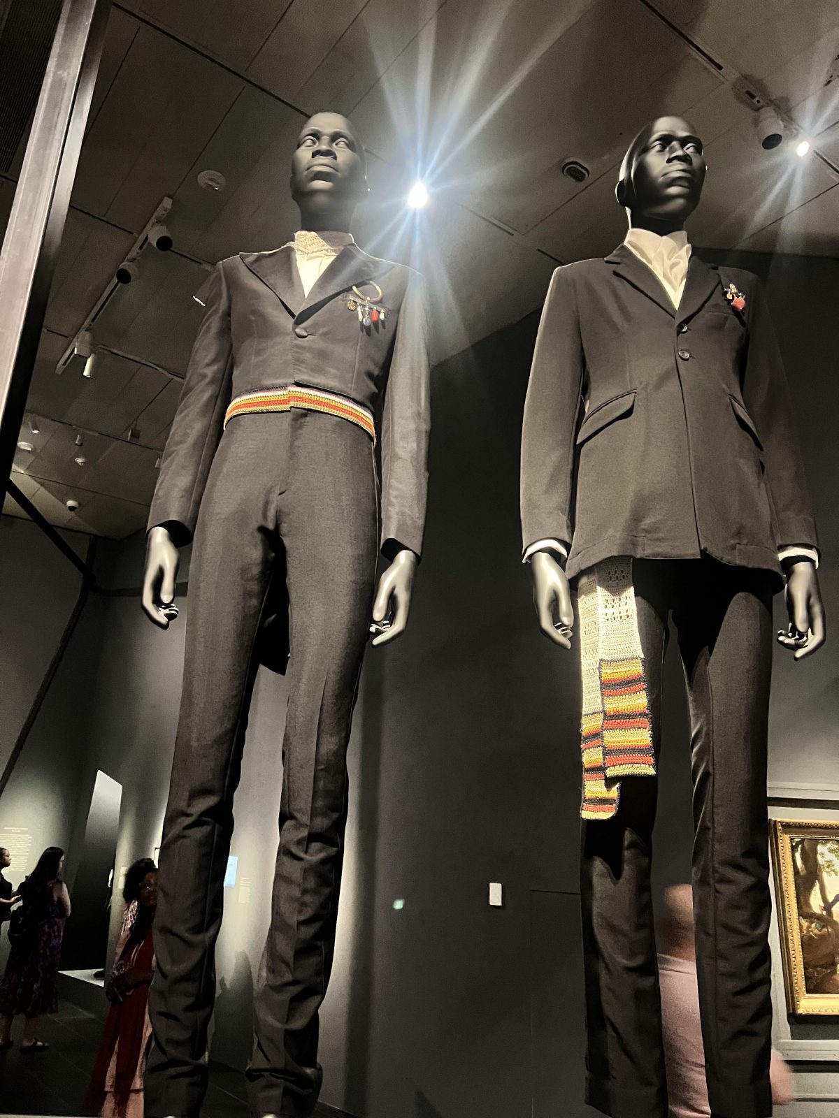 Two tall mannequins dressed in tailored black suits stand in a museum exhibition. One suit features a colorful woven sash at the waist and a brooch, while the other has a long multicolored knit sash hanging from the pocket. Visitors are seen in the background observing the exhibit.