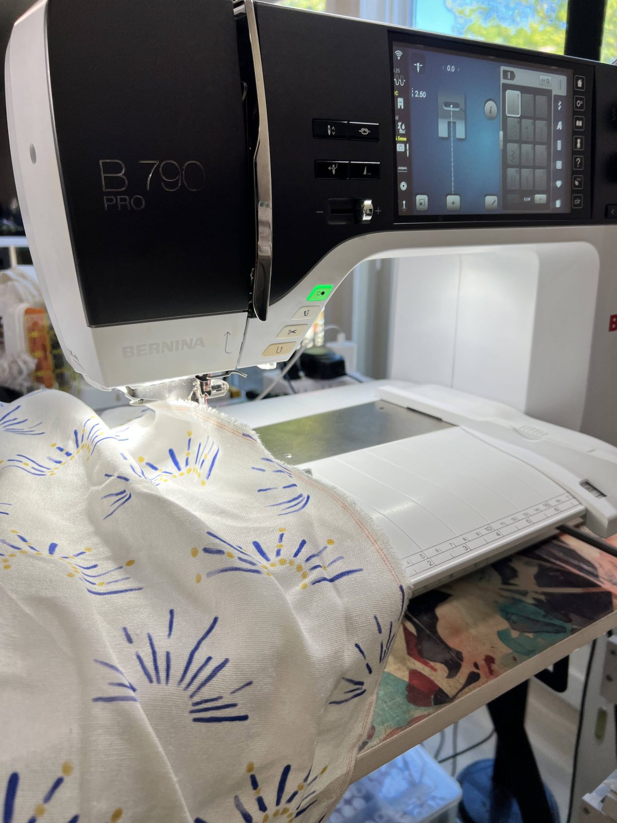 Close-up of a BERNINA B 790 PRO sewing machine stitching white fabric with a blue and gold suns. The machine screen is lit, and the fabric is being guided under the presser foot.