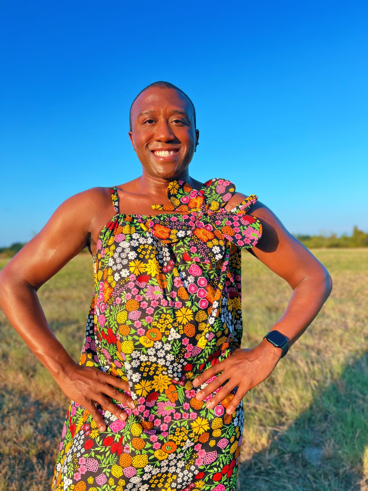 Close-up of a person wearing a colorful handmade floral Ankara maxi dress with thin straps and an oversized matching fabric flower at the shoulder.