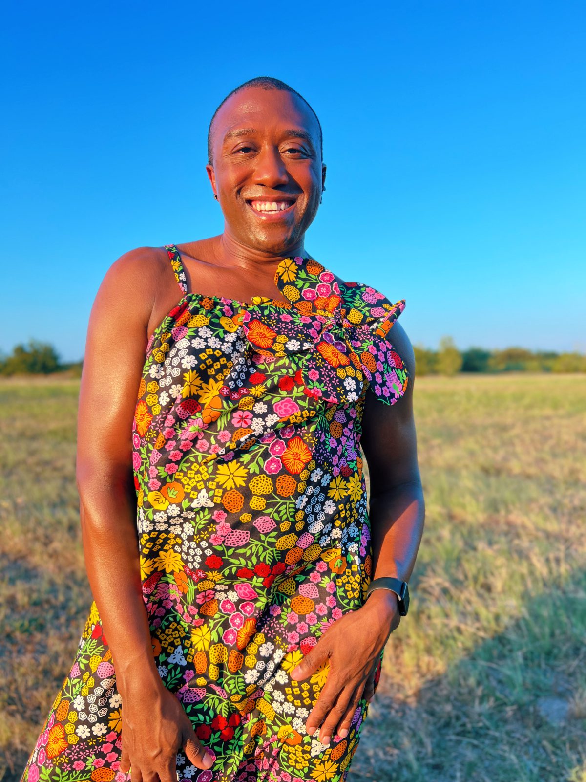 Close-up of a smiling person wearing a colorful floral Ankara maxi dress with thin straps and an oversized matching fabric flower tied at the shoulder.