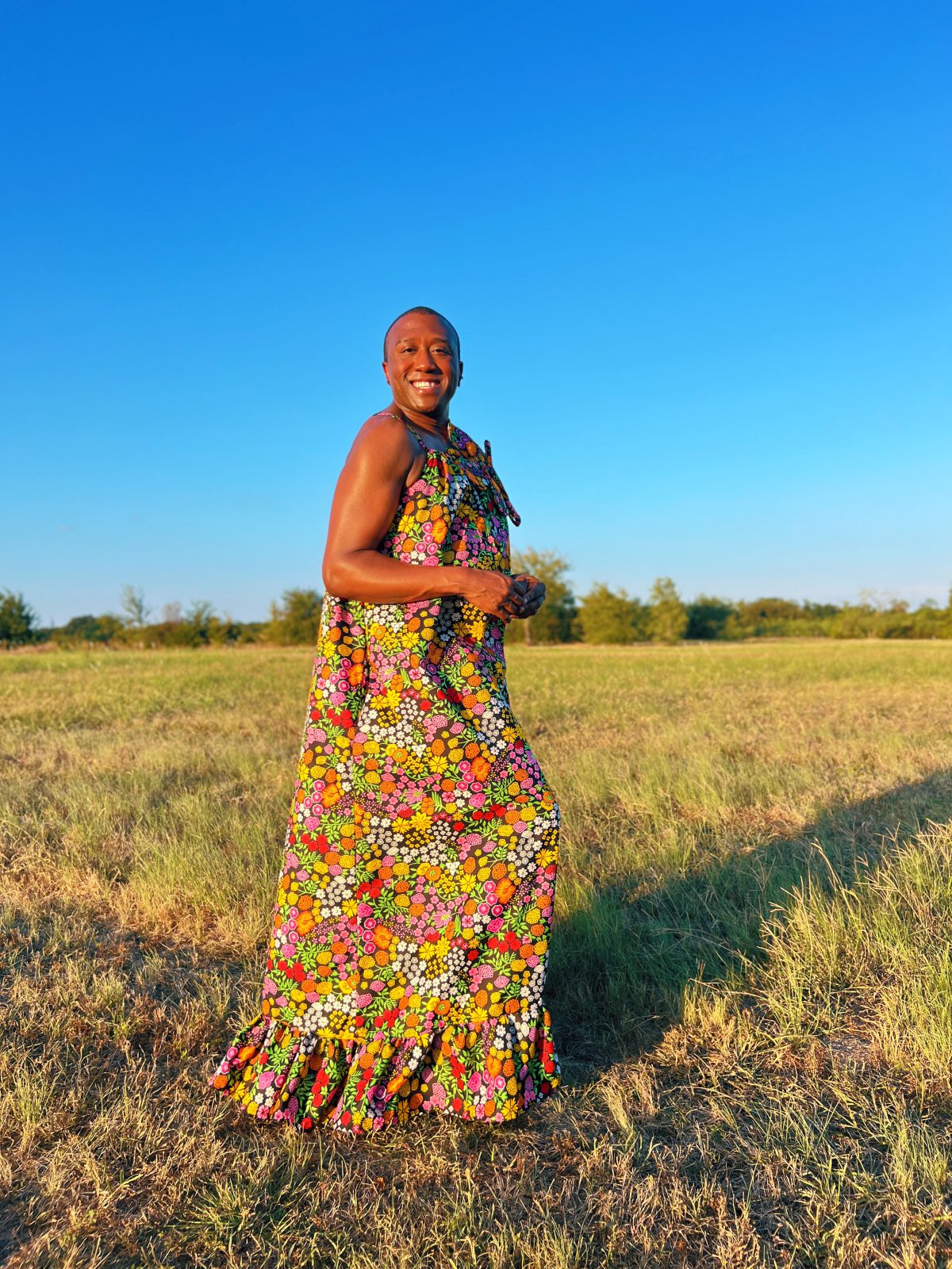 Smiling person wearing a colorful floral Ankara maxi dress with an oversized matching fabric flower at the neckline, photographed outdoors in a grassy field.