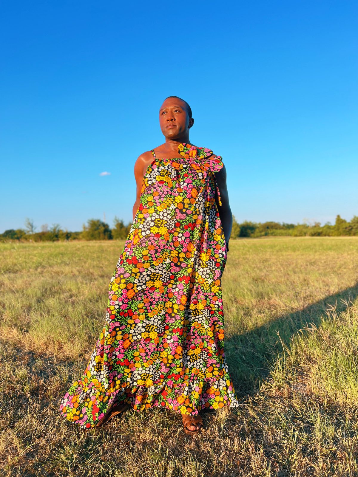 Smiling person wearing a colorful floral Ankara maxi dress with an oversized matching fabric flower at the neckline, photographed outdoors in a grassy field.