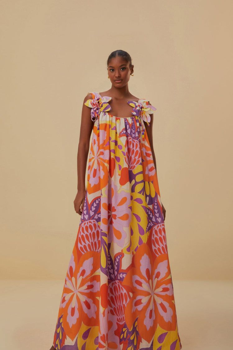 Model wearing a colorful Farm Rio maxi dress with a flowing silhouette, bold floral and abstract prints in orange, purple, yellow, and pink, and oversized floral shoulder details.