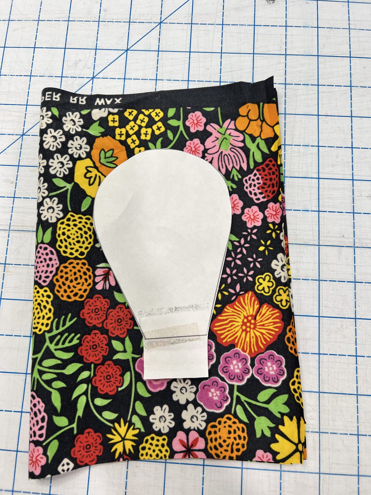 Large paper flower petal pattern piece placed on colorful floral Ankara fabric with pink, yellow, orange, red, white, and green flowers on a black background.