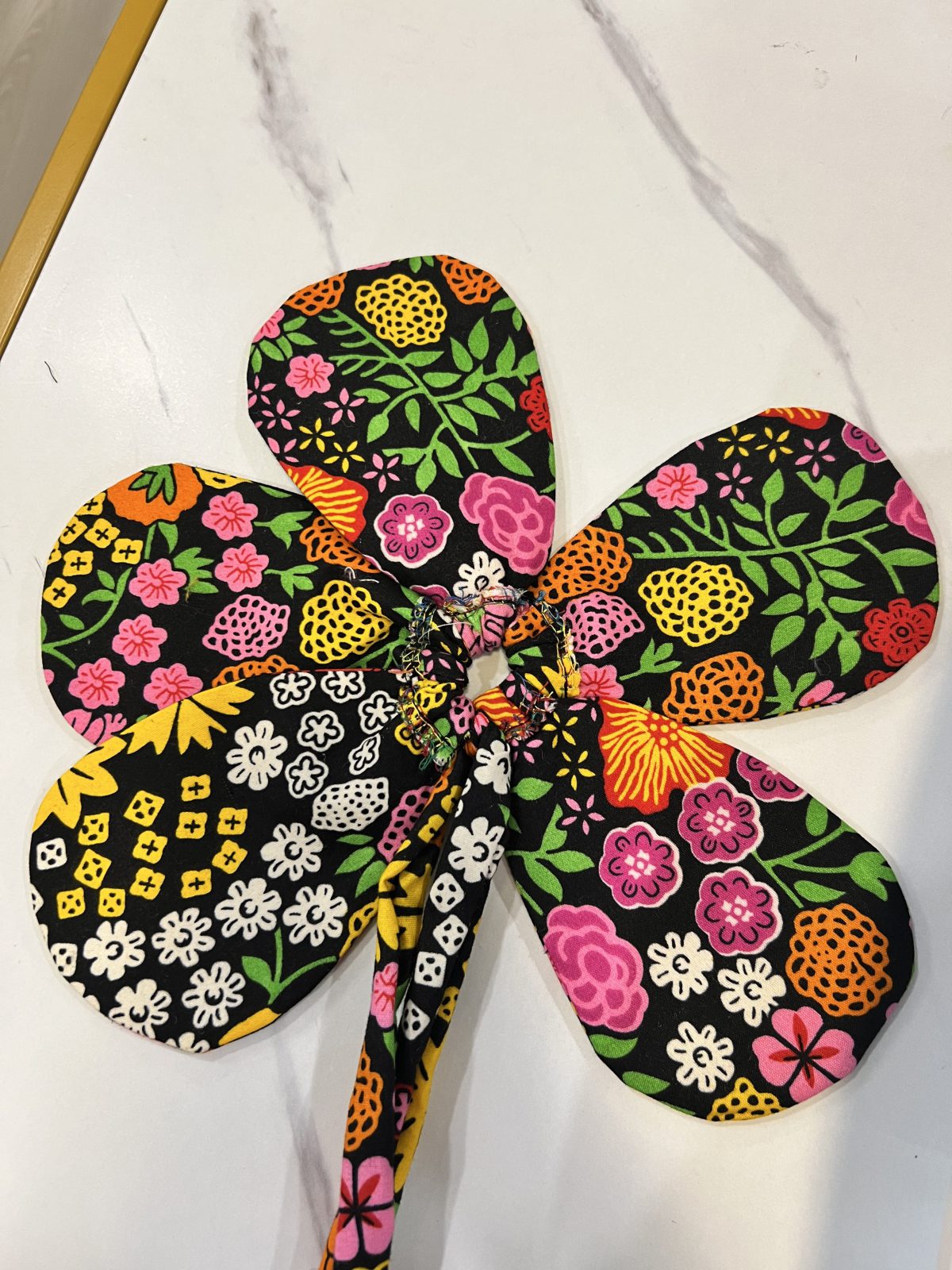 Oversized five-petal fabric flower handmade from vibrant floral Ankara fabric with pink, yellow, orange, red, white, and green botanical motifs on a black background.