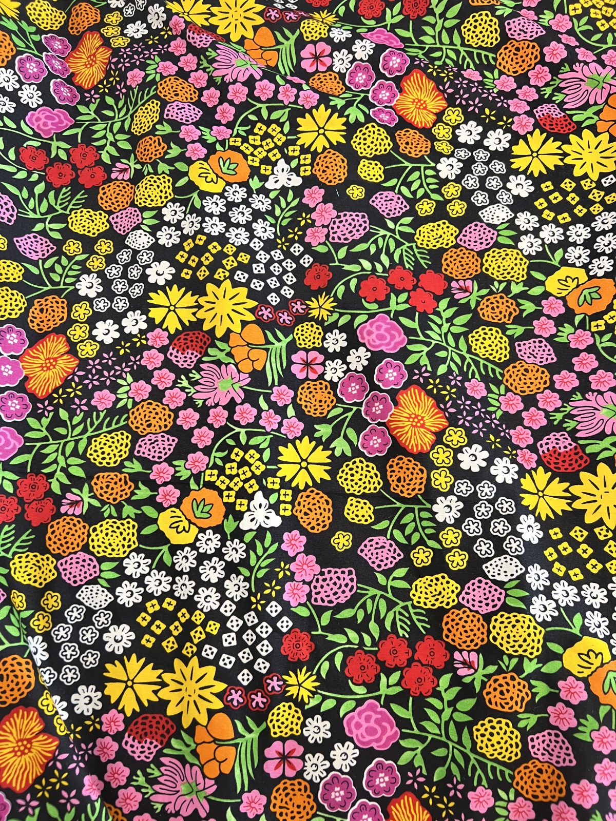 Bold Ankara fabric featuring a dense hand-drawn floral print with pink, yellow, orange, red, white, and green flowers and foliage against a black background.