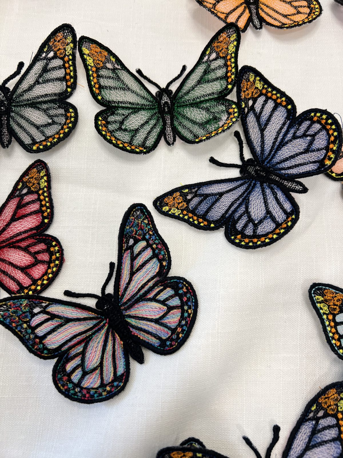 Close-up of colorful freestanding embroidered butterflies with black stitched outlines and detailed green, blue, pink, orange, and multicolored wings arranged on white fabric.