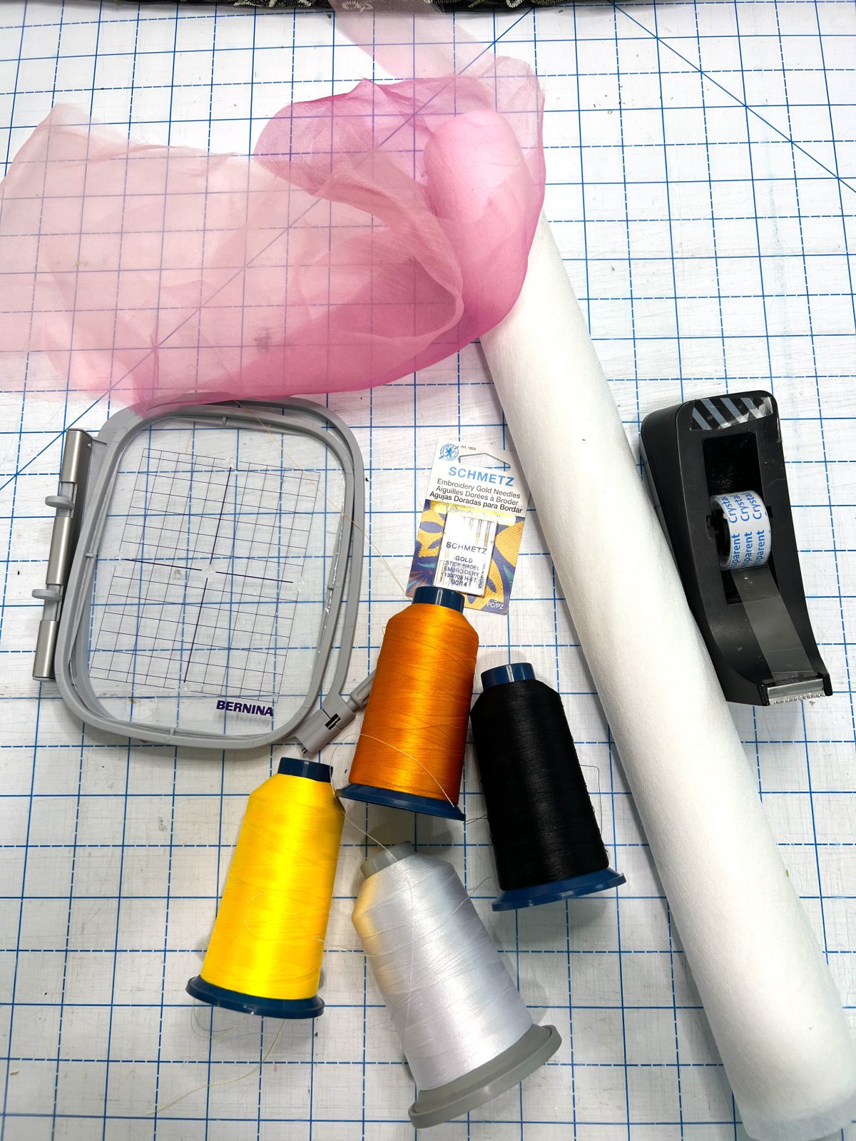 Machine embroidery supplies including pink tulle, white stabilizer, an embroidery hoop, embroidery needles, and colorful spools of thread arranged on a cutting mat.